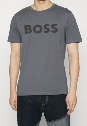 Mężczyzna noszący szary T-shirt z krótkim rękawem, na którym "BOSS" jest wydrukowane dużymi czarnymi literami na piersi, oraz ciemne dwukolorowe dżinsy.
