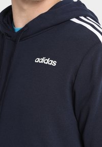 Tmavě modrá mikina Adidas z měkké tkaniny, zdobená bílými pruhy na ramenech a malým logem na hrudi.