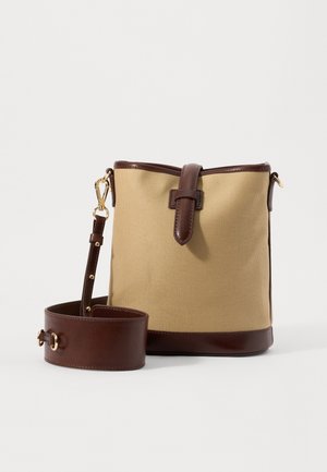 Sac seau en toile beige avec garniture en cuir marron, sangle à boucle, et large bandoulière ajustable avec ferrures dorées, posé sur une surface blanche.