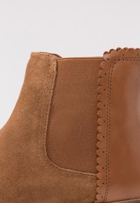 Bottine en suédine marron avec panneaux élastiques sur les côtés, détail de bordure en dentelle, et accents en cuir lisse. Présente un tissu texturé et des coutures.