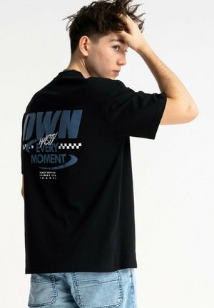 Młody mężczyzna o krótkich ciemnych włosach, ubrany w czarną koszulkę z nadrukiem i jasnoniebieskie dżinsy, dotykający głowy, stojący bokiem na białym tle.