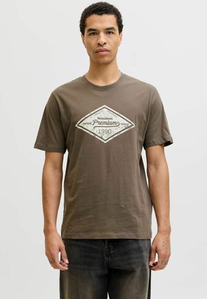 Mężczyzna w brązowej koszulce z białym vintage napisem "Jack & Jones Premium 1990", stojący na tle jasnego tła.