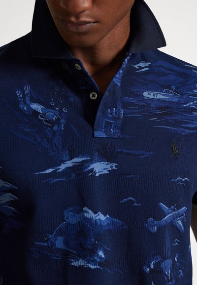 Vyras dėvi tamsiai mėlyną polo marškinėlį su po vandeniu ir aviacijos tema iliustracijomis, įskaitant narą, valtį ir lėktuvą.