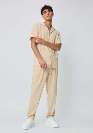 Homme debout portant une chemise beige à manches courtes avec col, un pantalon ample assorti, des baskets blanches et des colliers superposés sur un fond uni.