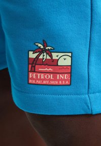 Tyrkysové kraťasy s červenou a krémovou nášivkou, na které je zobrazený palmový strom, slunce a vlny, označené nápisem "PETROL IND." v tučném písmu.