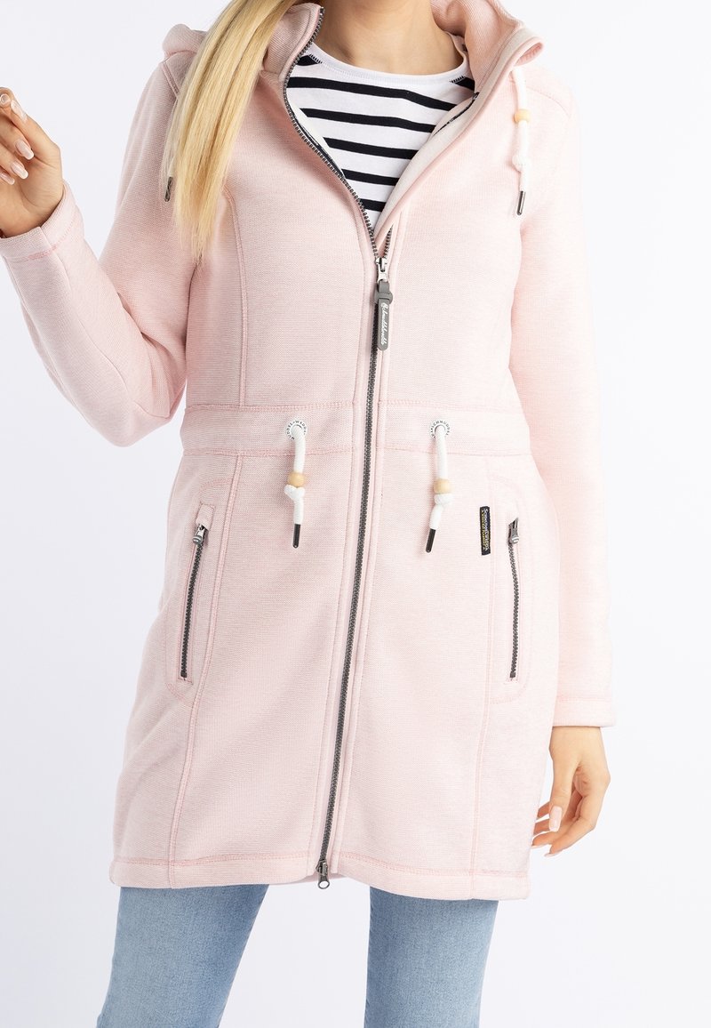 Femme portant une veste à capuche zippée rose clair avec des cordons blancs, un t-shirt rayé noir et blanc, et un jean bleu sur fond blanc.