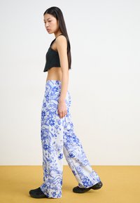 Un haut noir court associé à un pantalon ample bleu et blanc à motifs floraux et paysagers, porté avec des chaussures noires.