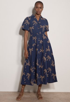 Donna che indossa un vestito midi blu navy con bottoni, stampe di palme color sabbia e maniche corte a sbuffo, in piedi contro una parete chiara e semplice.