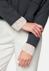 Grauer Pullover mit strukturierten, geschlungenen Ärmeln und cremefarbenen Spitzenmanschetten. Das Outfit wird mit weißen Hosen kombiniert.