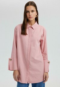 Chemise à rayures roses avec col, manches longues ornées de détails en bouton doré, et ourlets fendus. Tissu doux avec une coupe décontractée.