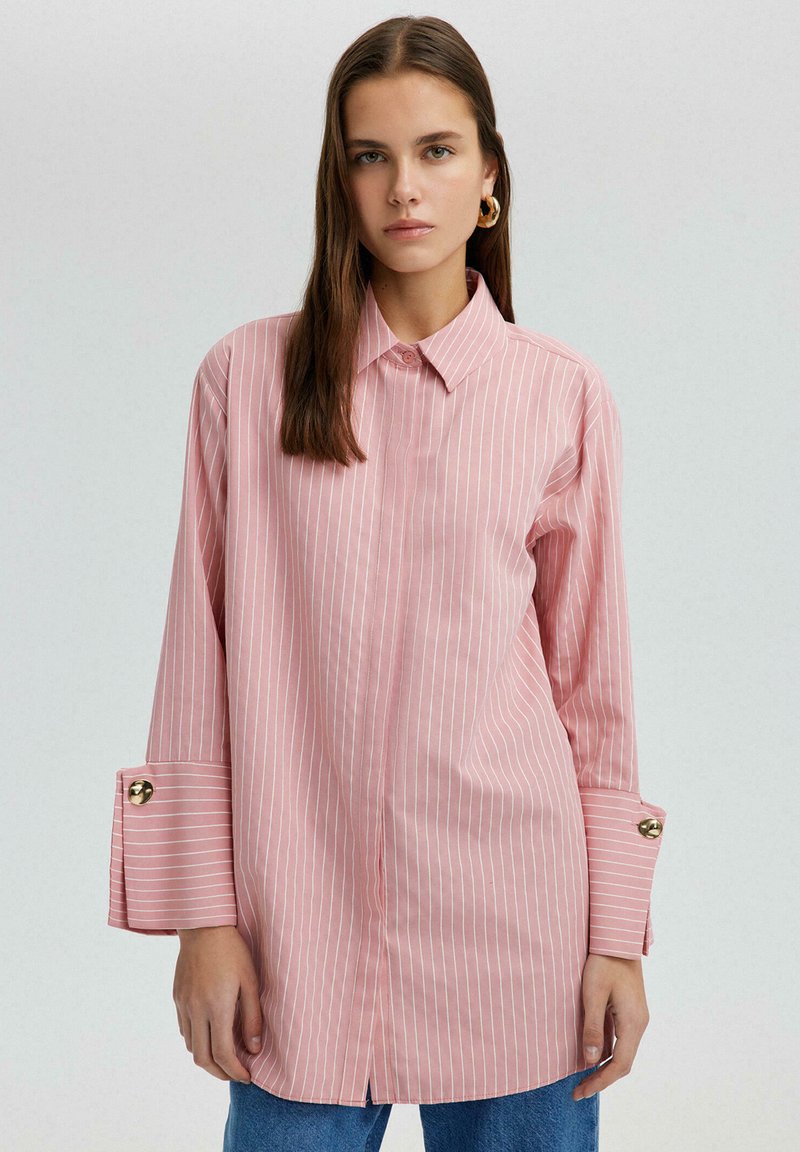 Chemise à rayures roses avec col, manches longues ornées de détails en bouton doré, et ourlets fendus. Tissu doux avec une coupe décontractée.