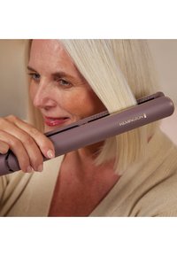 Lilla hårstyler med en slank design, som har en matt finish og gyldne detaljer. Brukeren fører den langs rette, lyse blonde hår.