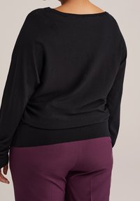 Černý svetr se širokým výstřihem a volným střihem, s dlouh ými rukávy. Kombinován s přiléhavými fialovými kalhotami, které mají hladkou strukturu.