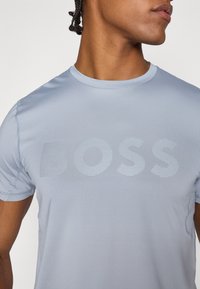 Világoskék sportpóló, körkivágással, rövid ujjal, és kifejező tónusos "BOSS" logóval csíkos részletekkel.