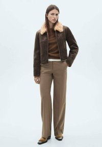 Geacă din suede maro cu guler din blană sintetică crem, purtată împreună cu un pulover tricotat maro și pantaloni largi de culoare maro deschis, având un design elegant.