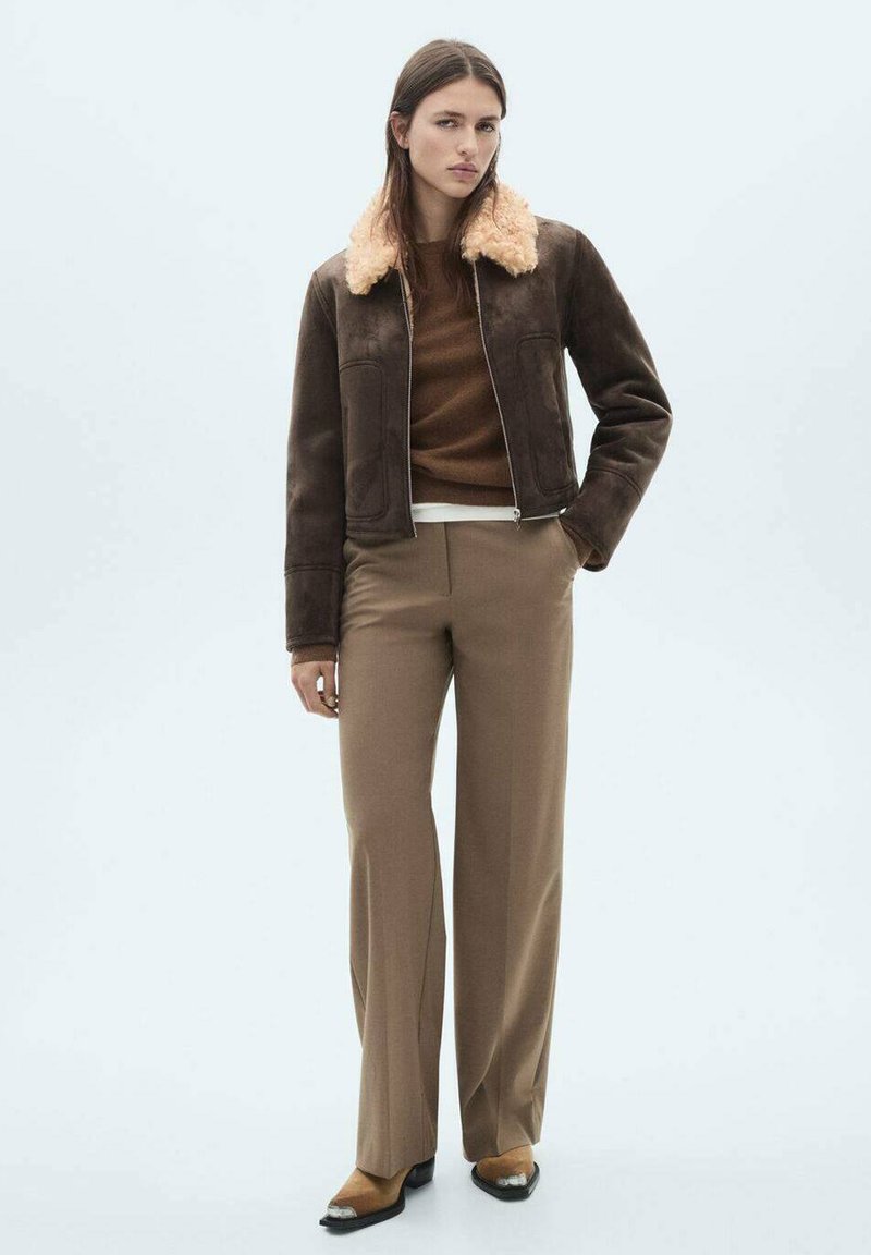 Geacă din suede maro cu guler din blană sintetică crem, purtată împreună cu un pulover tricotat maro și pantaloni largi de culoare maro deschis, având un design elegant.