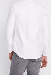 Camisa blanca de manga larga con puño abotonado, textura suave y costura en el centro de la espalda, combinada con pantalones grises que presentan un diseño de costura en los bolsillos.