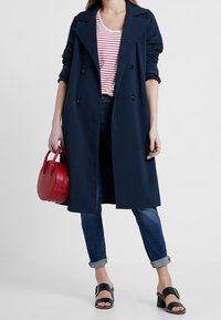 Gabardina navy de doble botonadura, combinada con un bolso rojo, camiseta a rayas, jeans remangados y sandalias negras con tiras al tobillo.