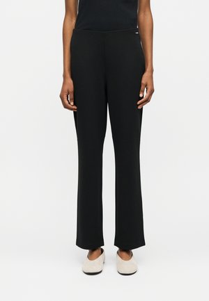 Osoba ubrana w czarne spodnie o prostych nogawkach i beżowe buty wsuwane, stojąca na tle jednolitego białego tła.