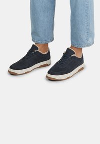 Jūras spēku zilo zamšādas sporta apavu augšdaļa ar teksturētu virsmu, baltā vidusdaļa un brūnā gumijas zole. Iekšpuse apkārt kaklam ir mīksts mākslīgais kažoks.