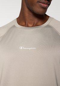 Μπεζ αθλητική μπλούζα με κοντά μανίκια και στρογγυλή λαιμόκοψη, με το λογότυπο Champion σε λευκό, κατασκευασμένη από υλικό με ομαλή, υφασμάτινη υφή.