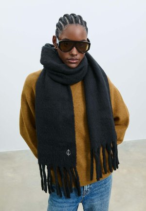 Zwarte wollen sjaal met franje aan de uiteinden, gedragen over een bruine trui. Bevat een subtiele logodetail. Gladde textuur, verlengde vorm voor warmte.