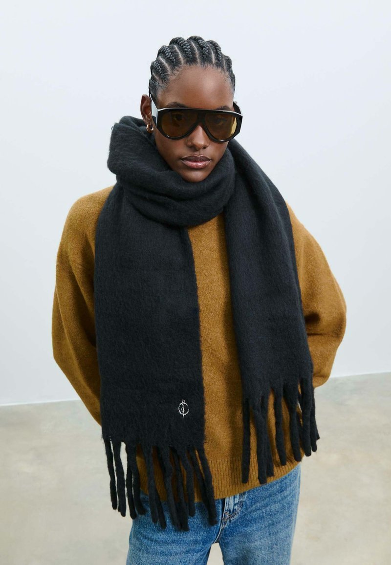 Sort uldhalstørklæde med frynser i enderne, båret over en brun sweater. Har et subtilt logo-detalje. Glat tekstur, forlænget form for varme.