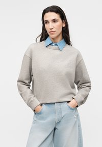 Mujer con cabello largo y oscuro, llevando una sudadera beige sobre una camisa azul con cuello y vaqueros azul claro rasgados, manos en los bolsillos, mirando ligeramente hacia la izquierda.