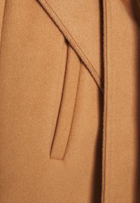 Manteau couleur camel en tissu doux et texturé. Présente un col à revers, des poches latérales et des coutures soignées le long des bords.