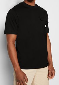 T-shirt en coton noir avec col rond, manches courtes et une poche poitrine. Comprend une petite étiquette logo sur la poche comme détail.