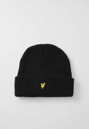 Bonnet en tricot noir avec une texture côtelée et un revers replié, arborant un petit logo brodé jaune d'un oiseau à l'avant.