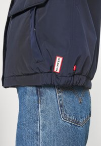 Marinblå Hunter-jacka med en röd logotyp över fickan på jeans av blå denim med orange sömmar.