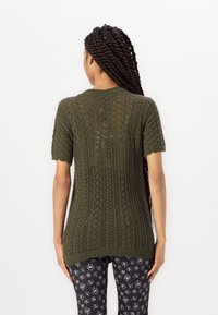 Frau mit langen, gedrehten Haaren, die einen dunkelgrünen, kurzärmeligen Strickpullover und schwarze, floral gemusterte Hosen trägt, steht mit dem Rücken zur Kamera.