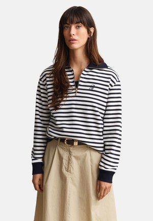 Frau trägt einen marineblau-weiß gestreiften Reißverschluss-Pullover mit schwarzen Bündchen und einen khakifarbenen, hoch taillierten Rock mit braunem Gürtel.