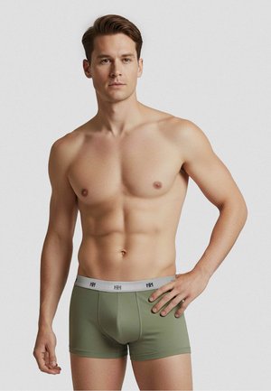 Atletische man staande met hand op de heup, draagt olijfgroene boxershorts met een grijze tailleband tegen een effen lichte achtergrond.