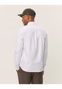 Camicia bianca con bottoni realizzata in tessuto dalla texture liscia, con uno spacco sul retro e maniche lunghe, abbinata a un cappellino verde.