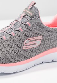 Szare siatkowe buty sportowe z różowymi akcentami, z teksturowaną powierzchnią, wyściełanym kołnierzem i białą podeszwą. Zawierają odblaskowy detal logo.