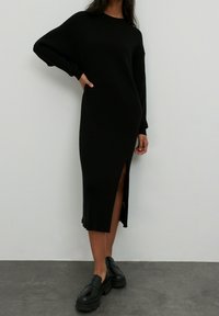 Robe noire en tricot côtelé avec des manches longues, fente sur le côté et coupe décontractée. Portée avec des chaussures noires épaisses, sur un fond uni.