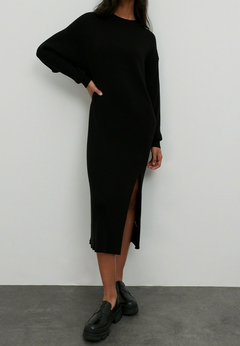 Robe noire en tricot côtelé avec des manches longues, fente sur le côté et coupe décontractée. Portée avec des chaussures noires épaisses, sur un fond uni.