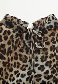 Blouse à col volanté imprimé léopard avec un détail de lien à nouer devant, présentant des taches marron, noires et beiges sur un tissu texturé.