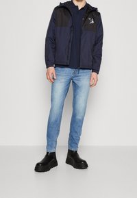 Granatowa rozpinana kurtka z czarnymi panelami, casualowa granatowa koszulka polo, jasnoniebieskie slim-fit dżinsy oraz czarne masywne buty.