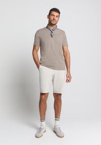 Beige T-shirt met korte mouwen, witte shorts, grijze sneakers. Blauw-witte scarf met patroon om de nek. Effen achtergrond in neutrale tint.