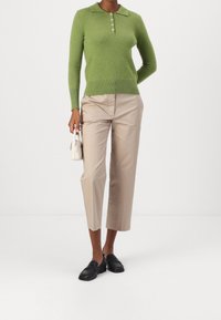 Kobieta w zielonym swetrze polo z długim rękawem, w beżowych spodniaczkach do kostek, w czarnych loafersach, trzymająca małą białą torebkę, stojąca na tle białej ściany.