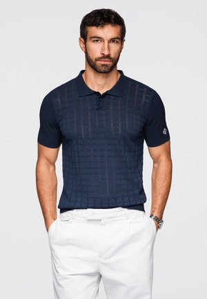 Uomo con barba che indossa una polo blu navy a maniche corte con motivo e pantaloni bianchi, in piedi con le mani in tasca su uno sfondo chiaro.