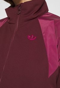Maroon athletic zip-up jacket with a high collar, featuring textured fabric and glossy pink side panels. Embroidered logo in pink on chest.