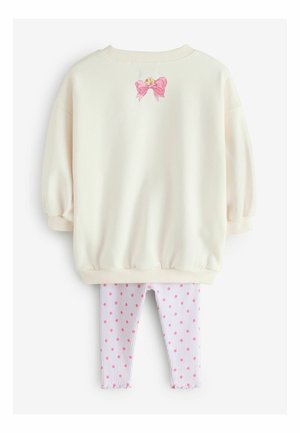 Blado kremowa bluza z bufiastymi rękawami i różową kokardą z tyłu, zestawiona z białymi legginsami w paski ozdobionymi różowymi kropkami.