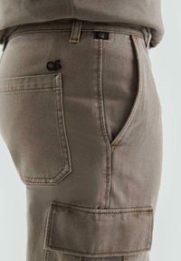 Beige cargobroek van stof, met een zijzak, gestikte accenten en een klein zwart logo label nabij de tailleband.