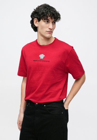 Κόκκινο βαμβακερό T-shirt με κοντά μανίκια και στρογγυλή λαιμόκοψη. Διαθέτει εκτυπωμένο λογότυπο Versace και γραφική λεπτομέρεια στο μπροστινό μέρος. Φοριέται με μαύρο παντελόνι.