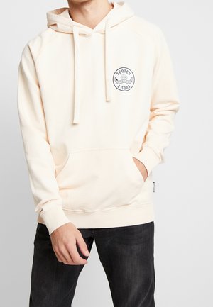 Osoba ubrana w jasnobeżową bluzę z kapturem z przednią kieszenią typu kangurka i logo "Scotch & Soda Netherlands" na lewej piersi, w połączeniu z czarnymi dżinsami.