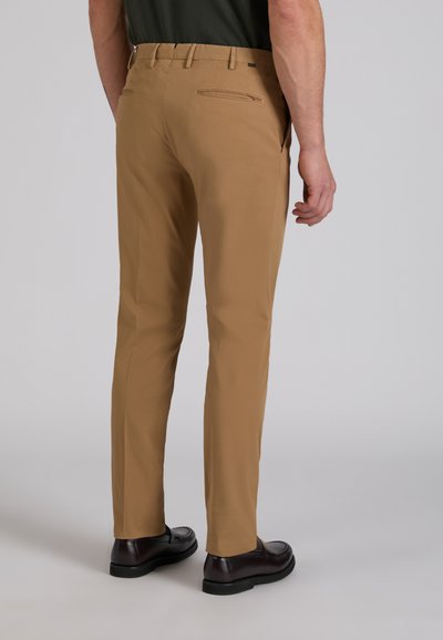 Mężczyzna w beżowych, dopasowanych spodniach i czarnych skórzanych mokasynach, stojący tyłem do kamery na jednolitym tle.